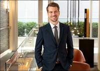 Professional headshot of man in navy suit, softly lit with city view.