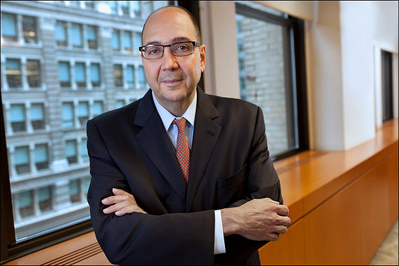 Corporate headshot of a CEO in suit and glasses, photographed in natural office light with NYC buildings visible through the windows.
