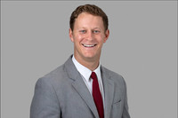 Professional headshot of smiling man in light gray suit on gray background.