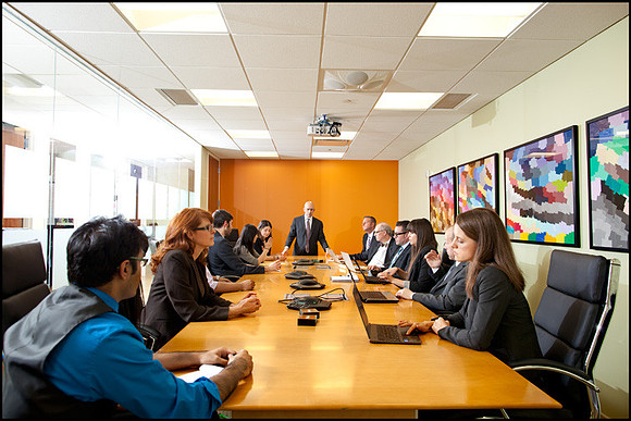 Executive team in a boardroom during a meeting, seated around a long table with laptops and notepads, NYC corporate setting.