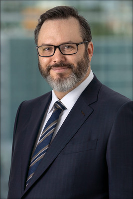 Corporate headshot of bearded man in glasses, wearing a suit and tie with blurred city skyline in background.