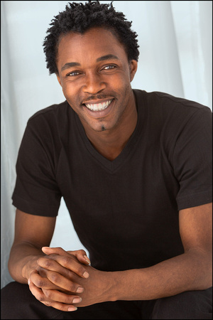 NYC actor headshot of young Black man in black tee, smiling with relaxed and confident energy.